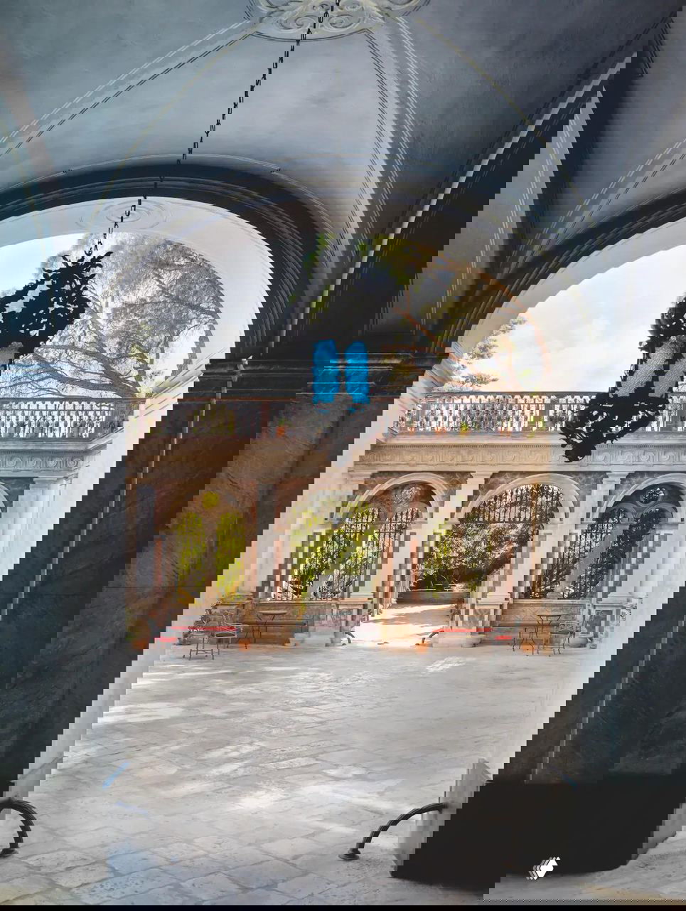 Арочный проём, ведущий во внутренний двор палаццо: висящий резной фонарь, колоннада и открытая терраса с видом на сад, подчёркивающие исторический характер объекта.