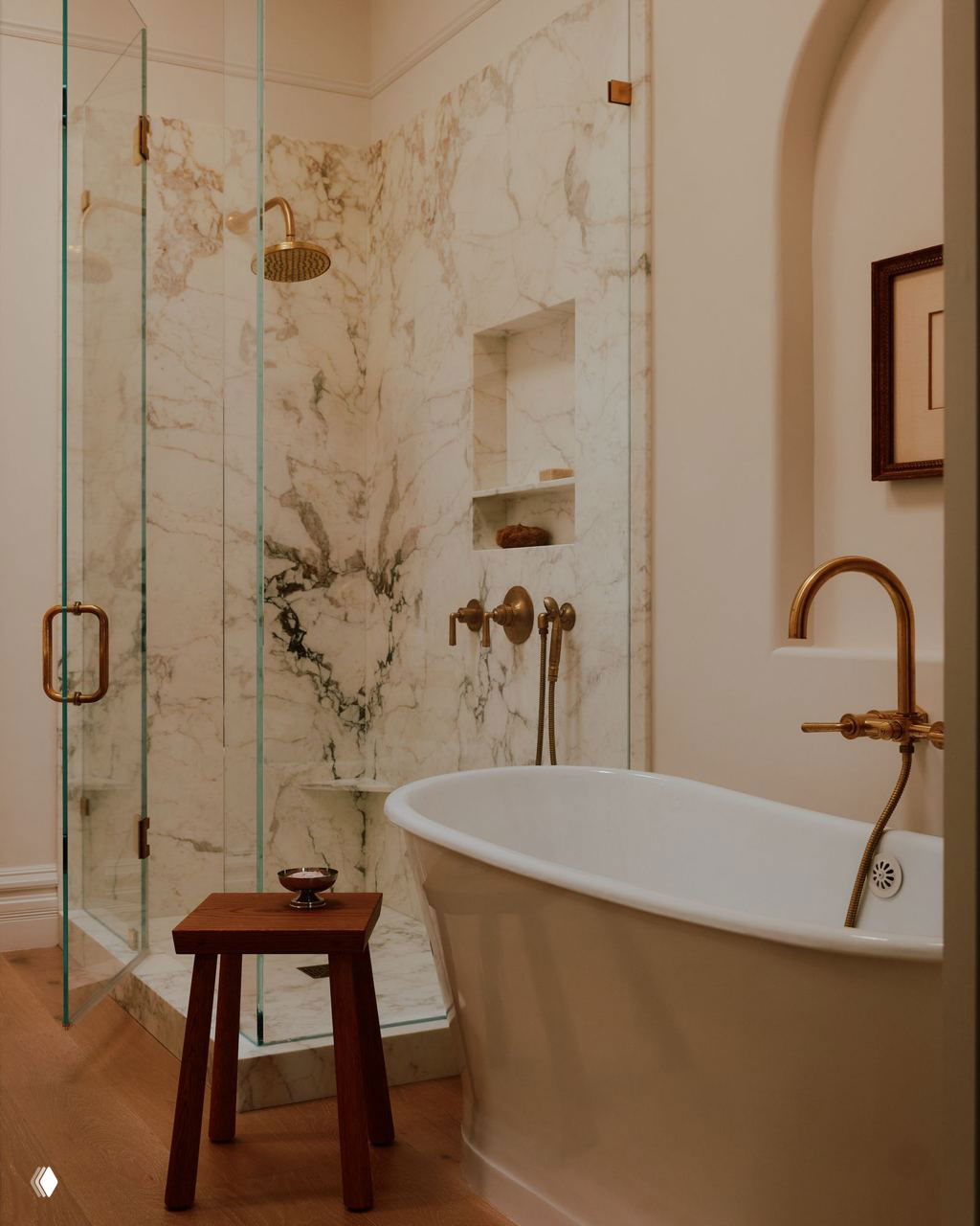 Ванная комната с мраморной душевой стеной, стеклянной перегородкой и отдельно стоящей белой ванной, бронзовые смесители и деревянный табурет для деталей.