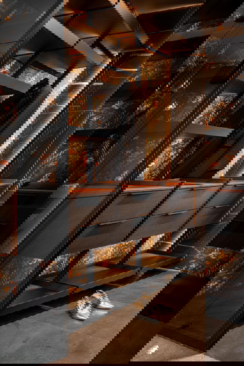 Стильная гардеробная с черной металлоконструкцией, темными фасадами, декоративными обоями с растительным орнаментом и подсвеченными полками для вещей и обуви.