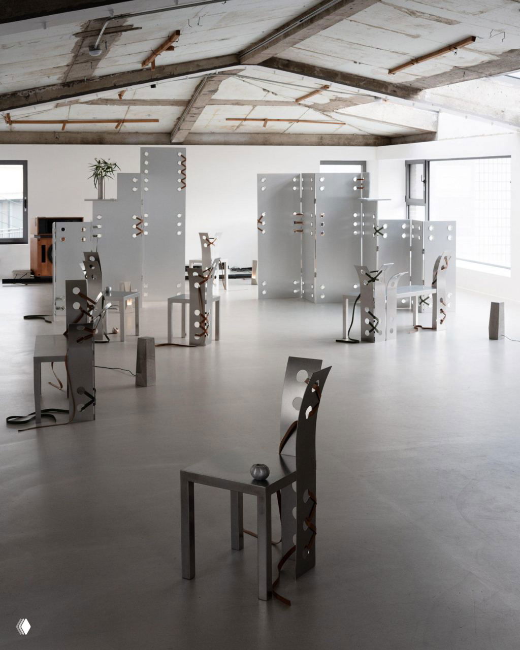 Общий вид выставки: несколько алюминиевых панелей и стульев с отверстиями и кожаными шнуровками, расставленных по залу с бетонным полом и высоким потолком.