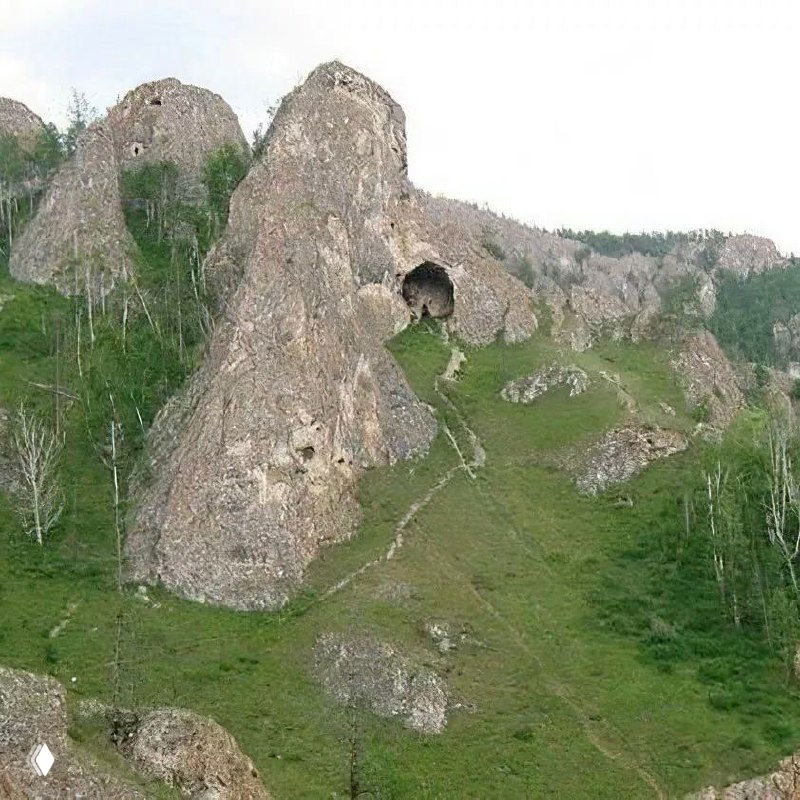 Скальный цирк в Кузнецком Алатау с крупным пещерным отверстием в середине уступа, пологие травянистые склоны и отдельные берёзки на склоне.