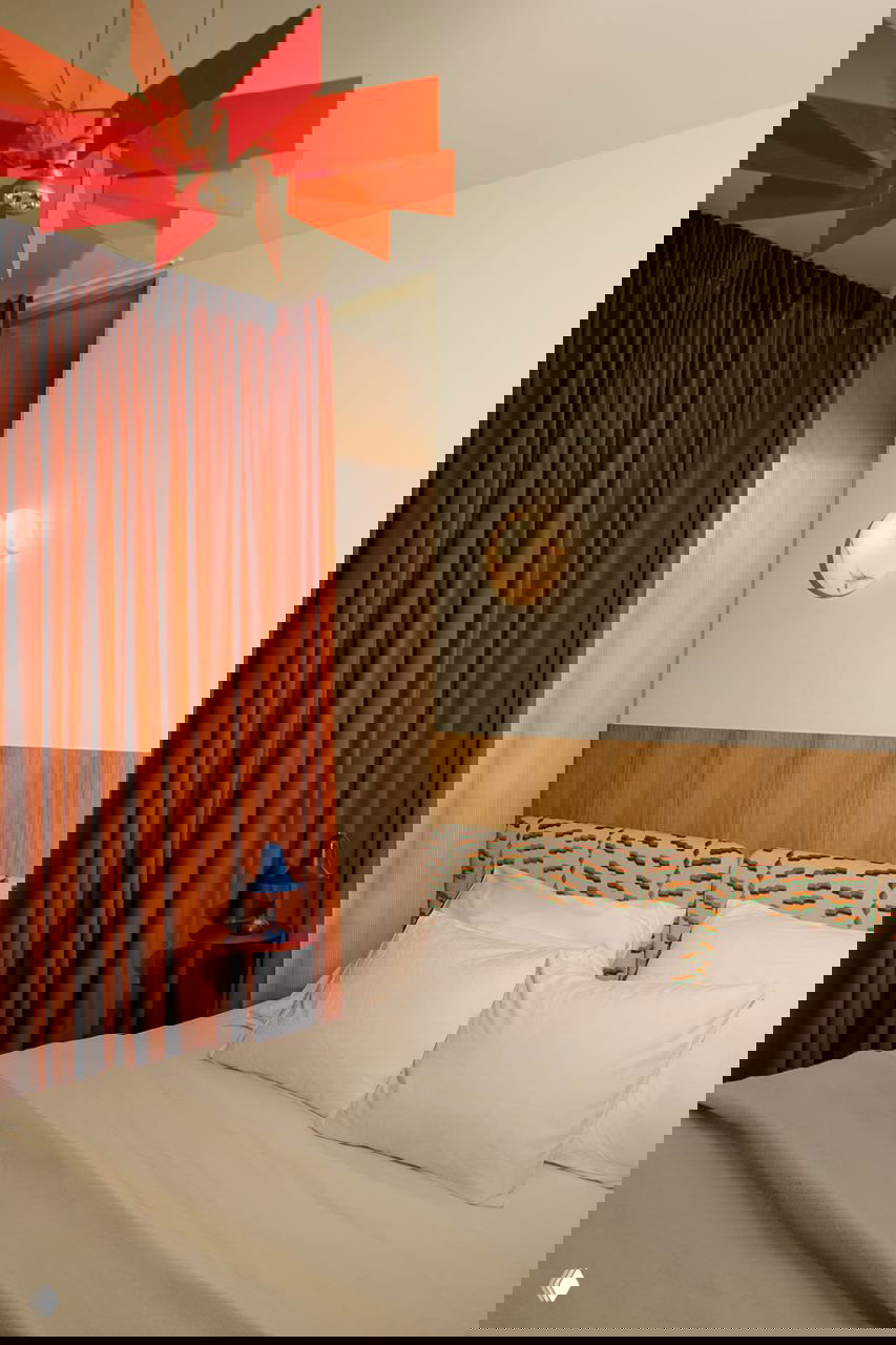 Еще один ракурс спальни: кровать с белым постельным бельем, крупные портьеры терракотового цвета и оригинальный подвесной светильник красного оттенка.