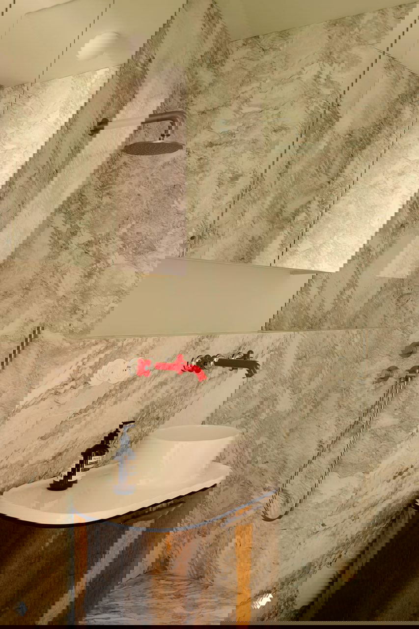 Ванная комната с мраморной облицовкой стен и душевой, небольшая овальная раковина на деревянной тумбе и яркий красный смеситель как дизайнерский акцент.