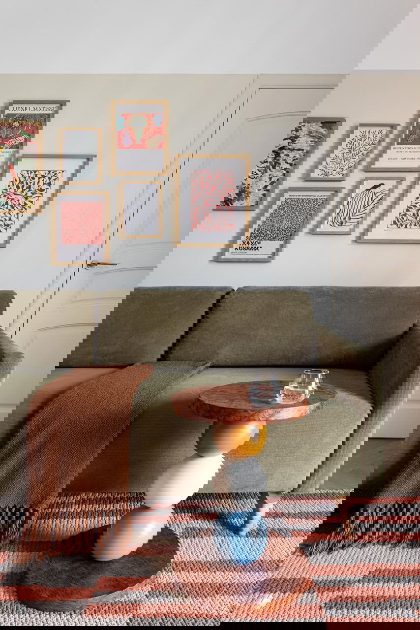 Гостиная с оливково‑зелёным диваном, небольшим круглым столиком‑скульптурой и настенной галереей рамок; тёплая, уютная композиция интерьера 50 м².