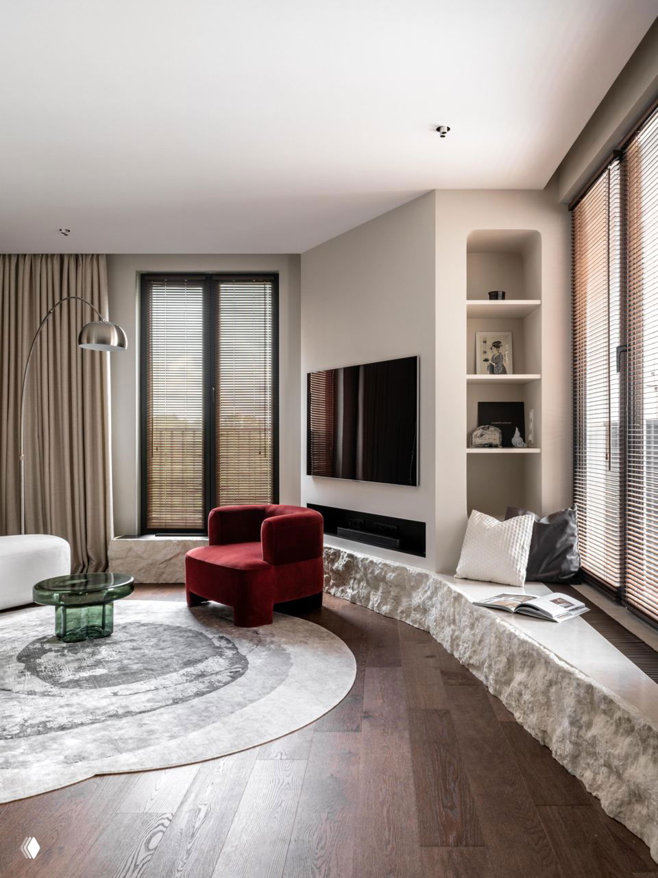 Гостиная в природной бежево‑коричневой гамме: круглый серо‑белый ковер, красное кресло, низкая каменная скамья у окна и тёмный деревянный пол.