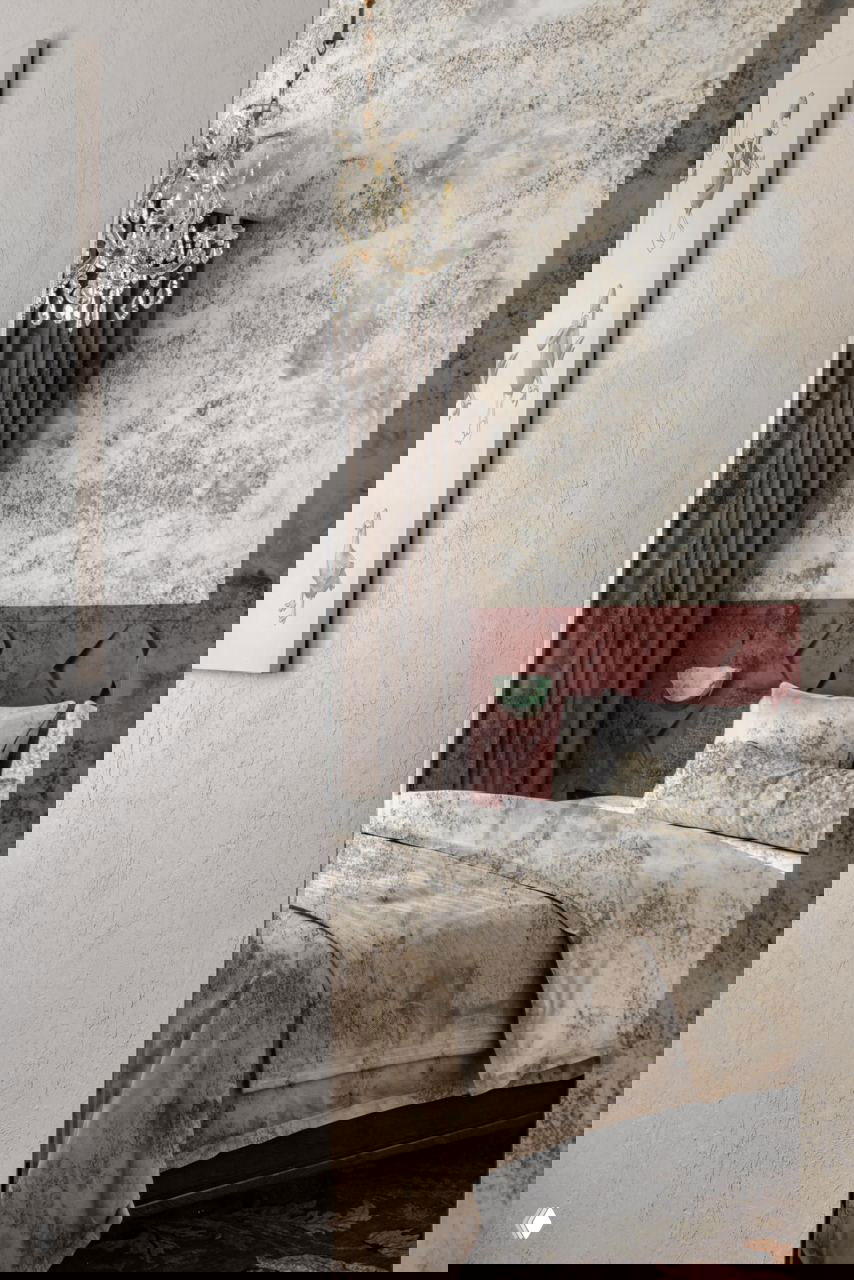 Спальня с текстурными стенами, бархатной изголовьем тёмно-терракотового оттенка и хрустальной люстрой, отражающейся в большом зеркале.