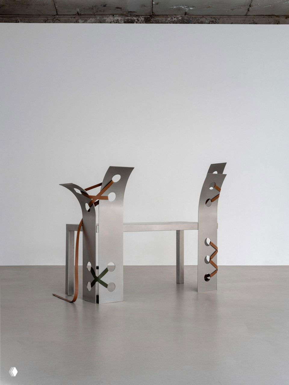 Минималистичная композиция из двух небольших скамеек/столов со стальными спинками и кожаными шнуровками на нейтральном фоне — акцент на форме и материале.