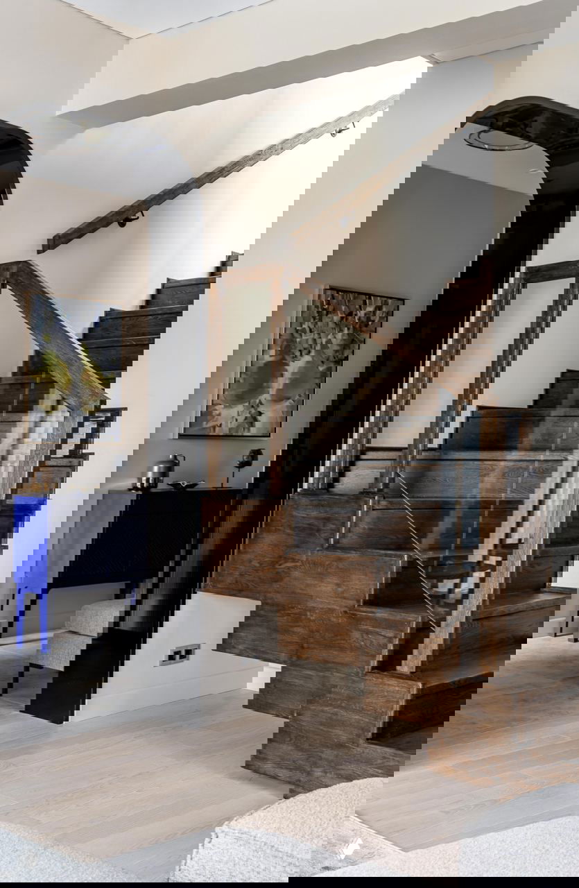 Прихожая с деревянной лестницей и стеклянным ограждением, светлый паркет и чёрная арка; вид подчёркивает контрастные акценты интерьера.