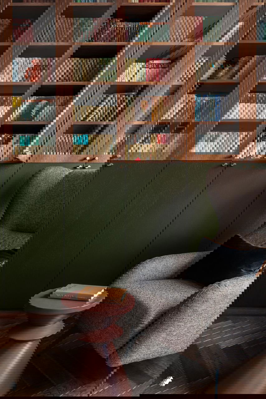 Уголок с книжным шкафом в тёмном дереве и низкими зелёными фасадами снизу, рядом уютное кресло и небольшой столик для чтения.