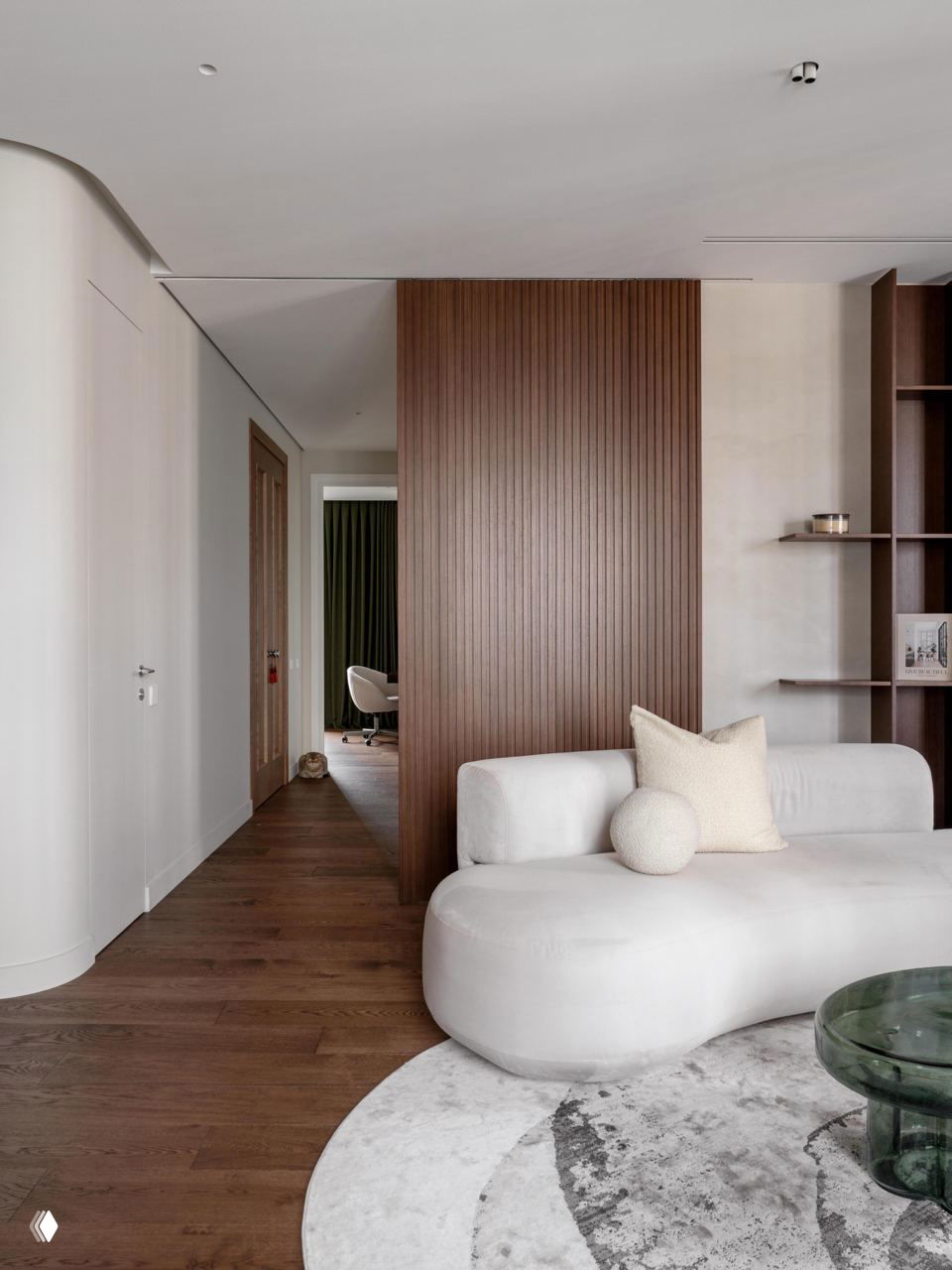 Мягкая белая серповидная софа и деревянная вертикальная панель-ширма, тёплый паркет и спокойные текстуры создают обнимающее пространство.