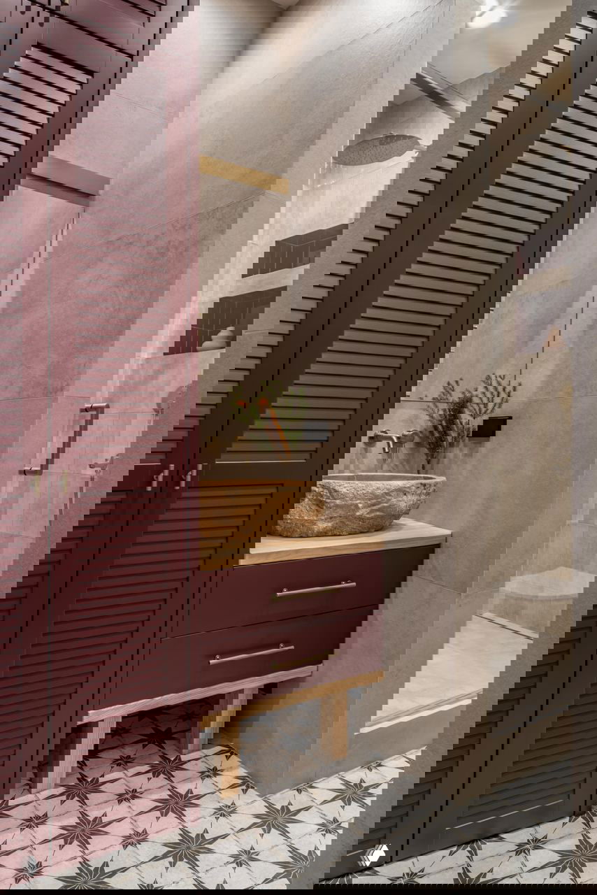 Ванная комната с серыми плитками, душевой кабиной, деревянной тумбой с каменной раковиной и бордовыми жалюзийными дверцами шкафа.