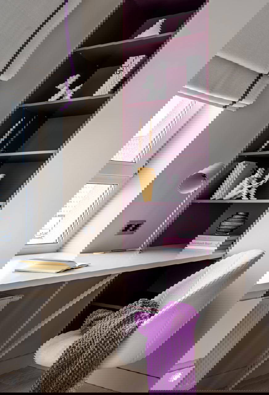 Рабочая зона у окна с белым столом и глубокими фиолетовыми полками, декоративные объекты и лампа создают уютный уголок для учёбы.