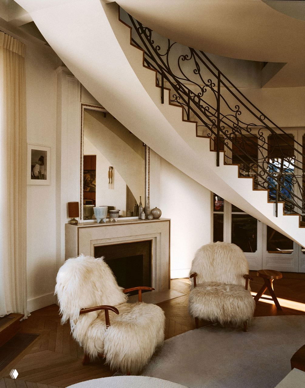 Гостиная под изогнутой лестницей с кованым перилом: два пушистых кресла, камин, зеркало и деревянный паркет в парижской квартире Изабель Станислас.