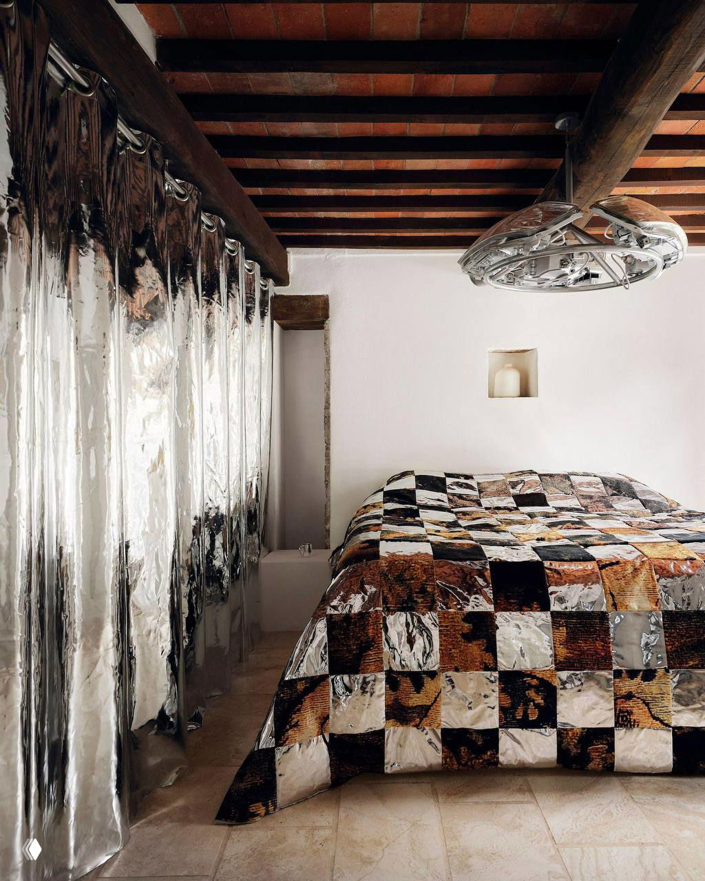 Спальня с лоскутным стеганым покрывалом и отражающими шторами; деревянные балки на потолке и минималистичная композиция предметов в интерьере.