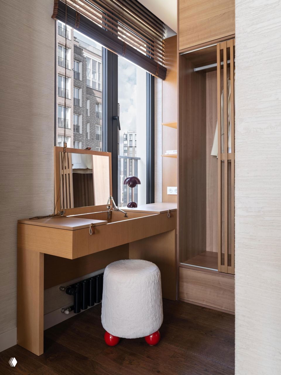 Туалетный столик у высоких окон: деревянная конструкция с откидными крышками, пушистый белый пуф на ножках и вид на город за стеклом.