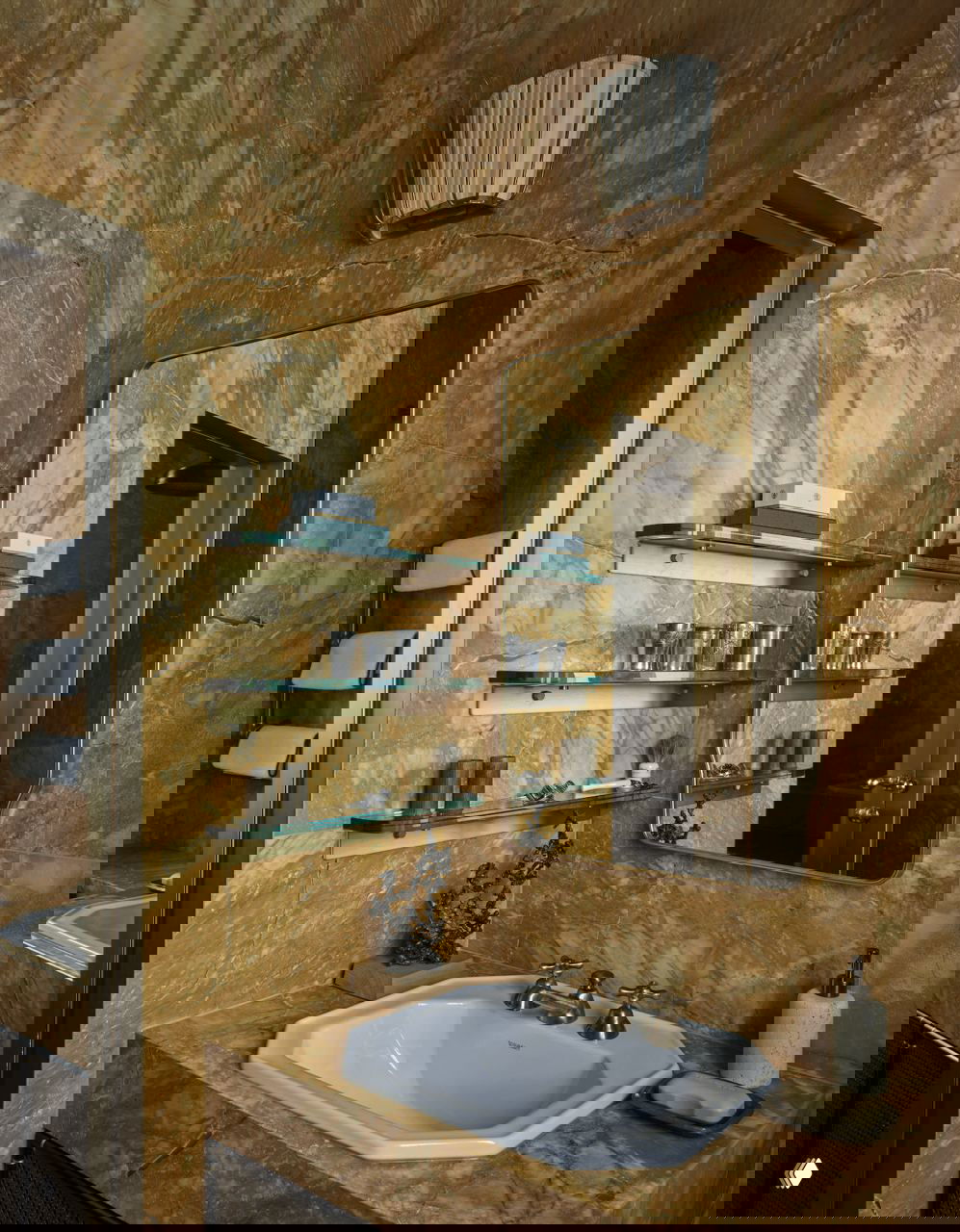 Ванная комната с мраморной облицовкой насыщенного золотистого тона: встроенная раковина, зеркало в латунной раме и стеклянные полочки с аксессуарами в ретро-стиле.