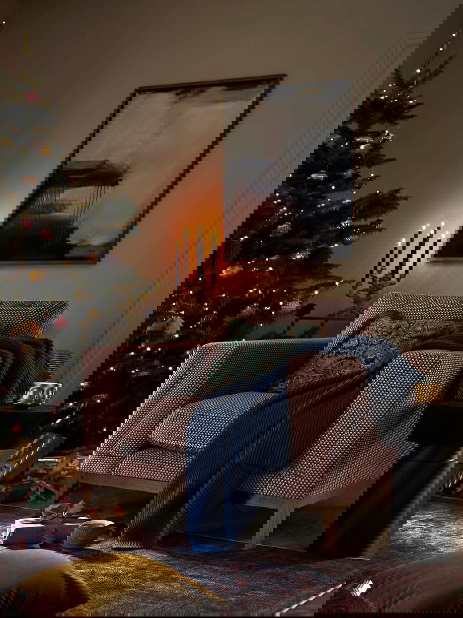 Уютный уголок гостиной с креслом и пледом на фоне распахнутой ёлки и свечей; тёплые оттенки и текстуры создают спокойную праздничную атмосферу для отдыха.