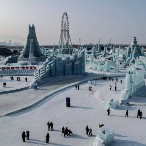 Широкая территория ледяного парка с колесом обозрения вдали, заснеженные дорожки и группы людей между ледяными скульптурами.