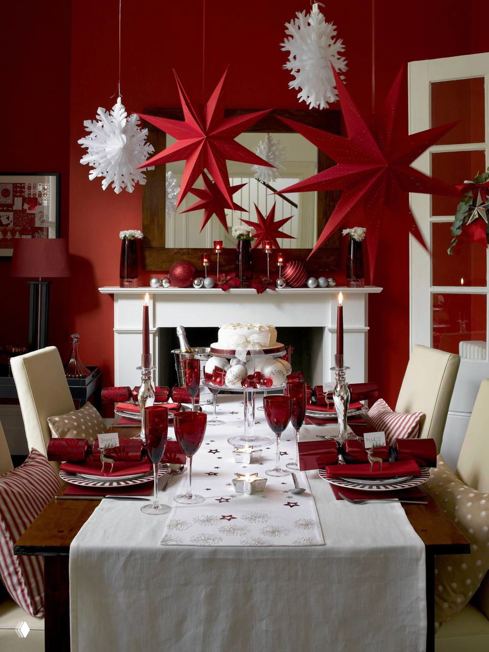 Яркая красная сервировка: висящие звёзды и снежинки, красные бокалы и свечи на столе перед камином — насыщенный праздничный интерьер в красных тонах.