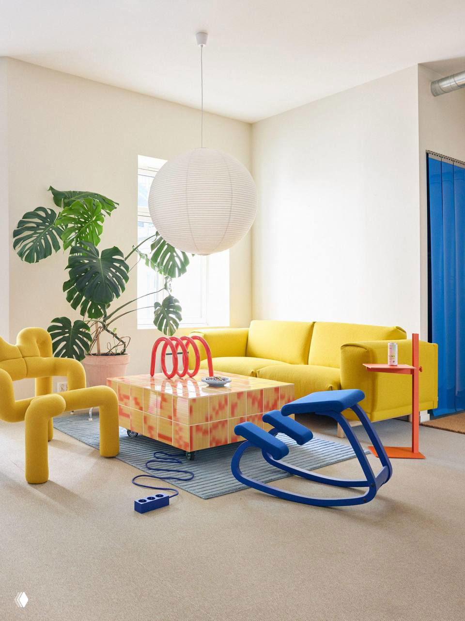 Яркая зона студии: желтый диван, скульптурные желтые кресла, синие качалки и квадратный журнальный столик в двухцветной плитке на фоне светлой стены и растения.
