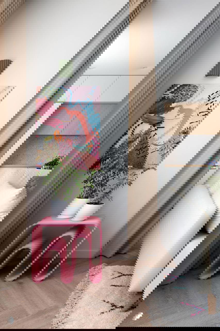 Интерьер: угол гостиной с белым диваном, бежевыми шторами, абстрактной картиной на стене и розовым декоративным столиком с растениями.