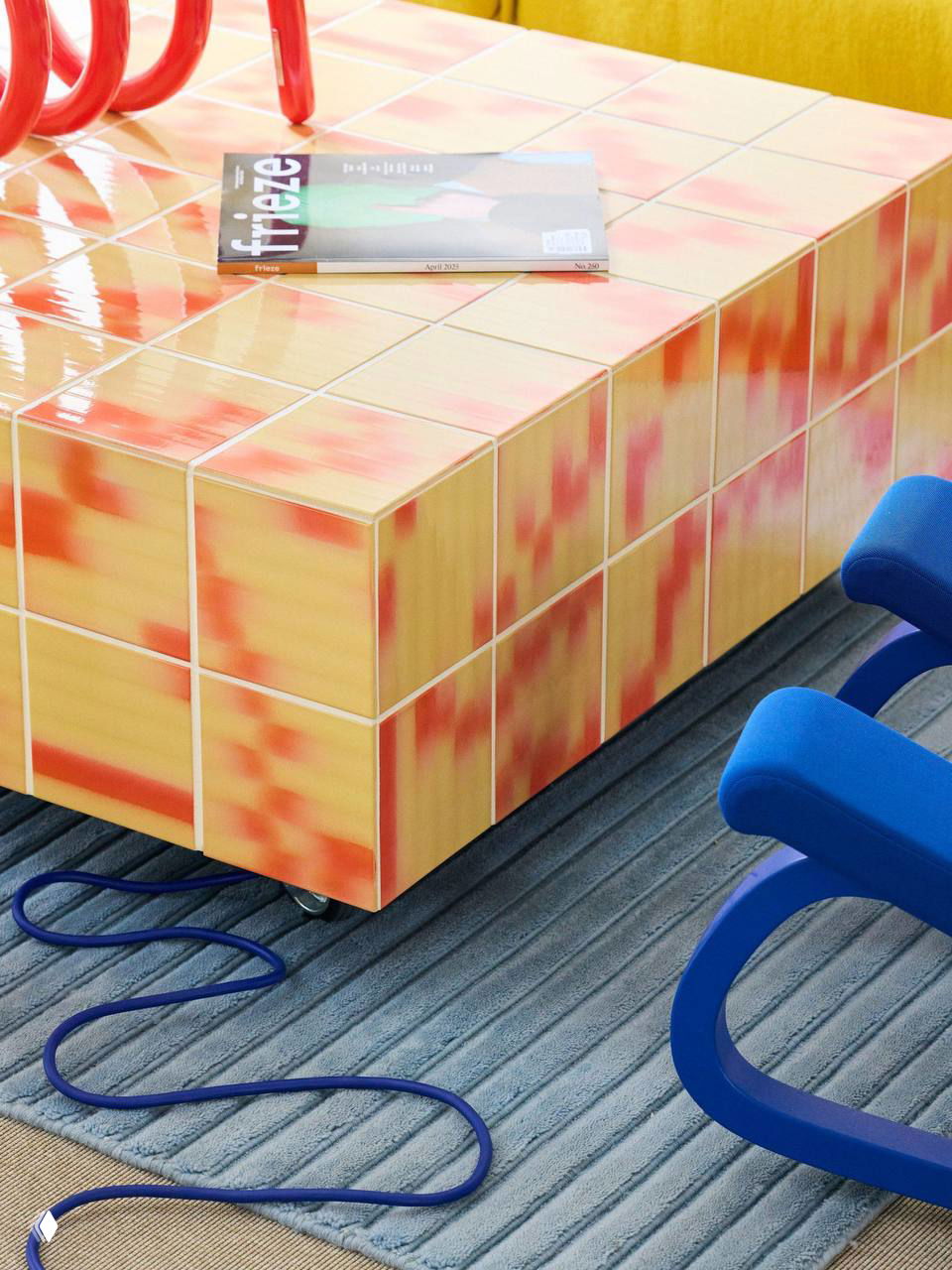 Крупный план журнального столика, покрытого двухцветной плиткой Palet: глянцевые квадратные плитки с желто-оранжево-красочным рисунком и деталями швов.