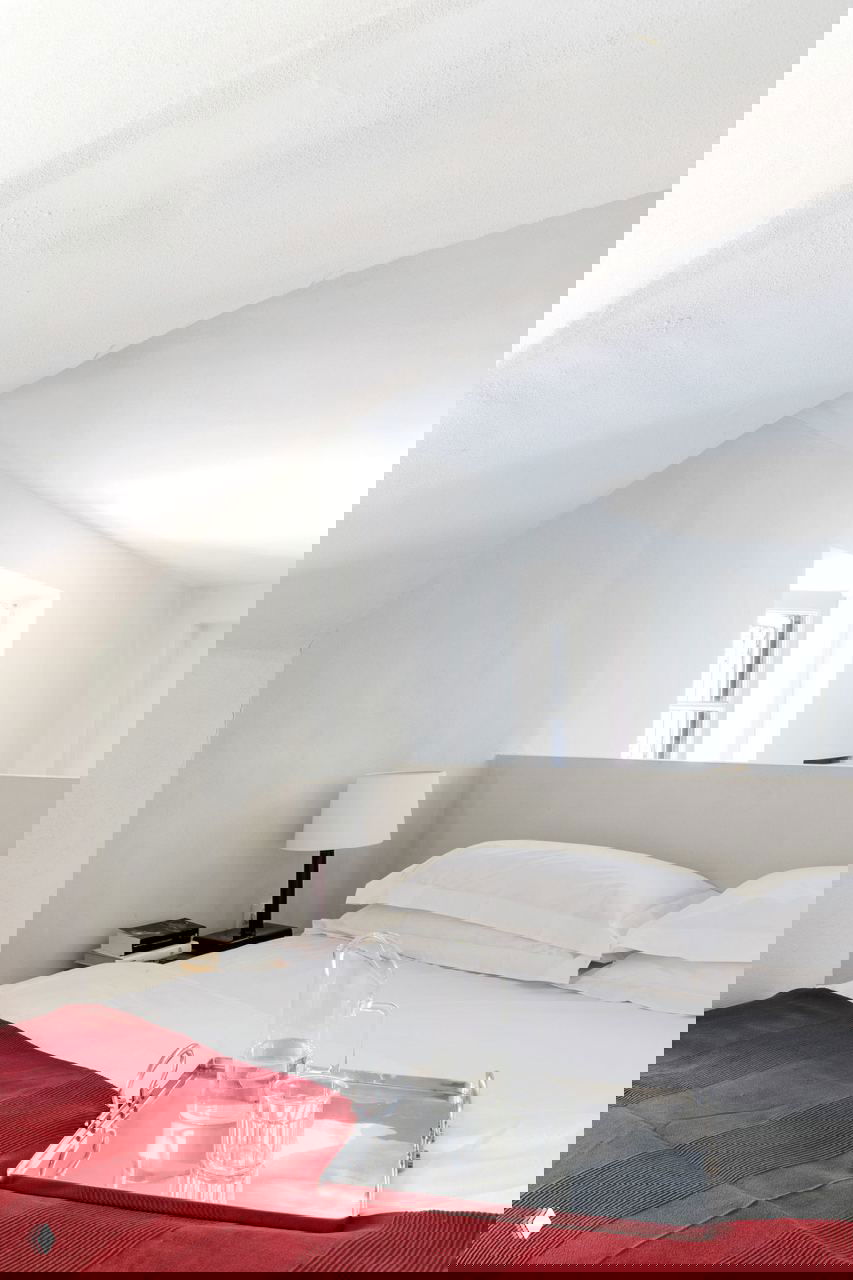 Спальная зона в белом интерьере микроквартиры: изогнутый потолок, простое белое постельное бельё и насыщенный красный плед с подносом на кровати.