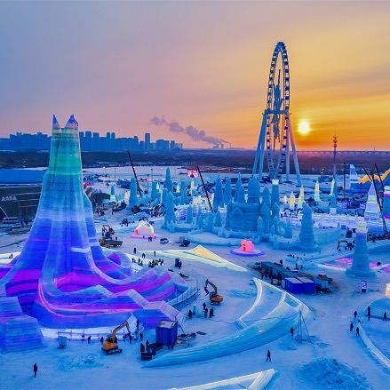 Закатный кадр с колесом обозрения и подсвеченными ледяными конструкциями на фоне горизонта, техника и люди на площадке фестиваля.