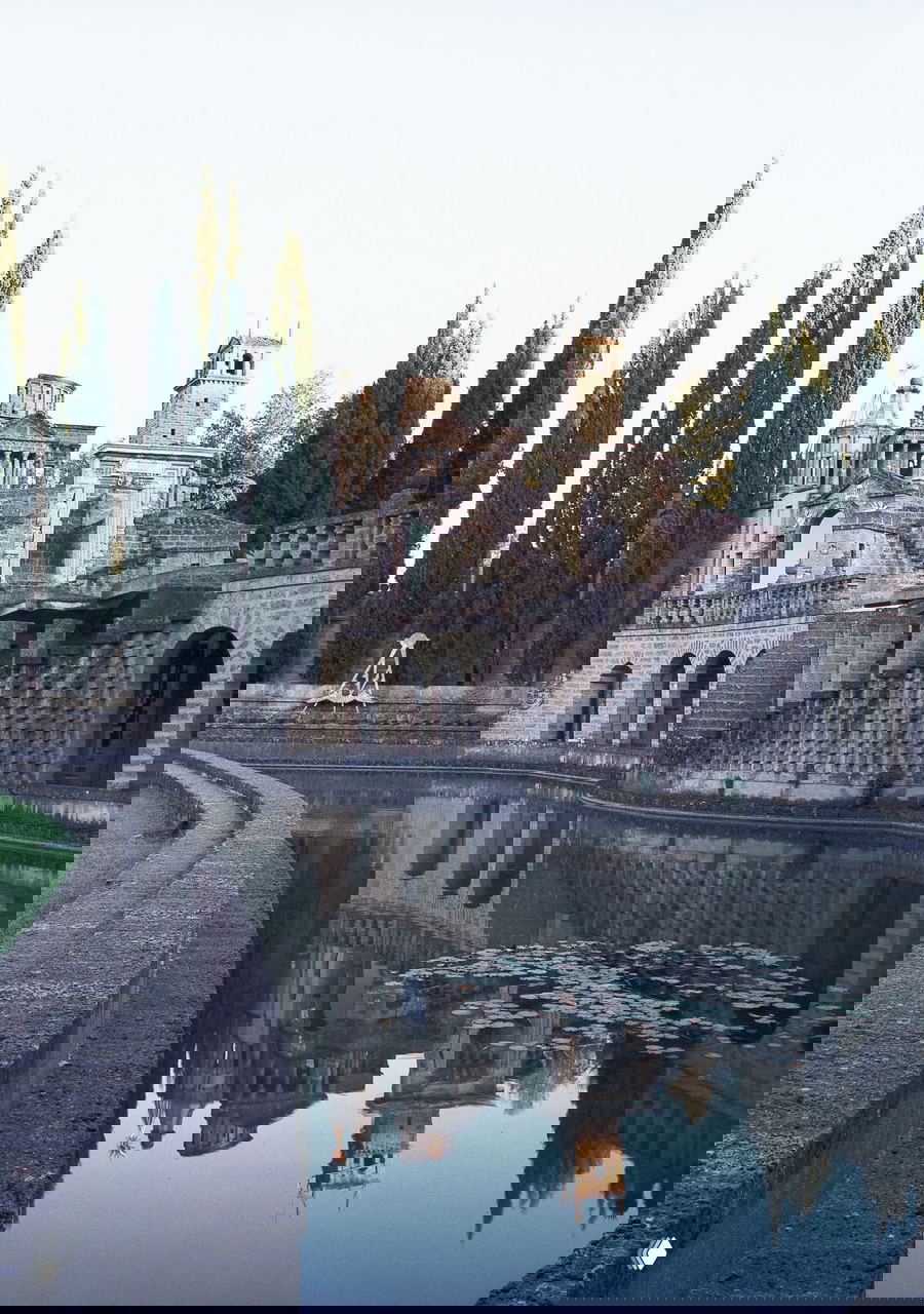 Панорамный вид на Ла Скарцуола: каменные террасы и арки у зеркального водоёма, окружённые кипарисами, с лёгким налётом времени.