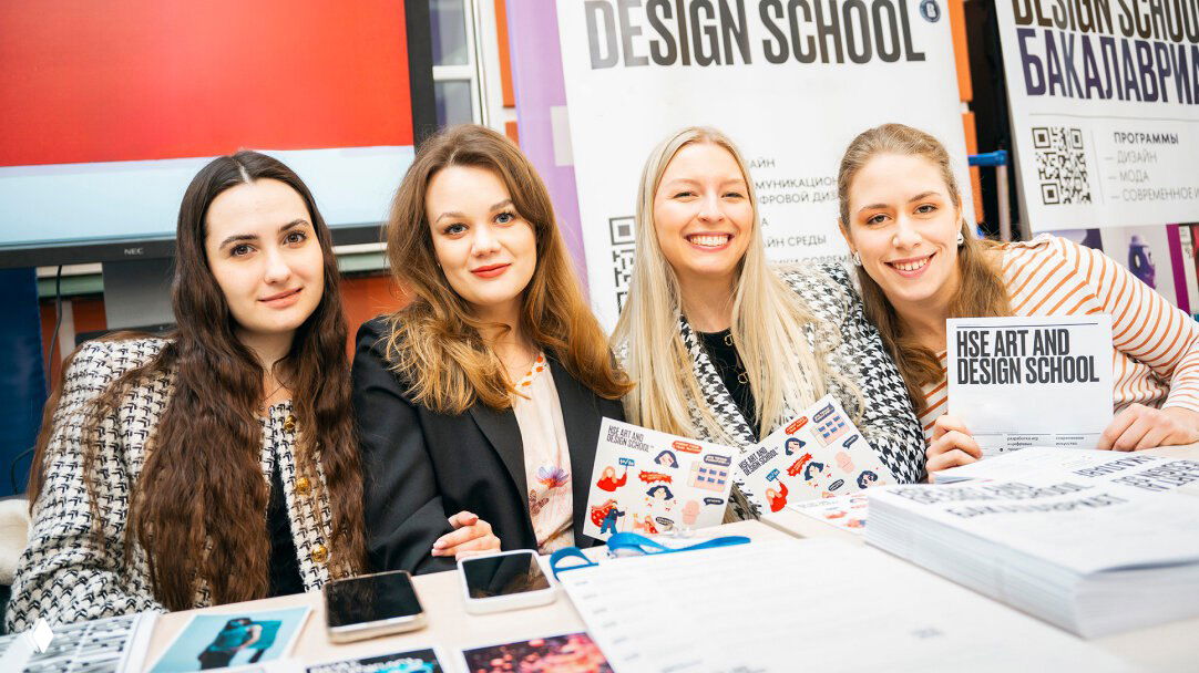 Улыбающиеся участницы у стола на стенде Школы дизайна НИУ ВШЭ: буклеты, телефоны и промо‑материалы на фоне баннера Design School.