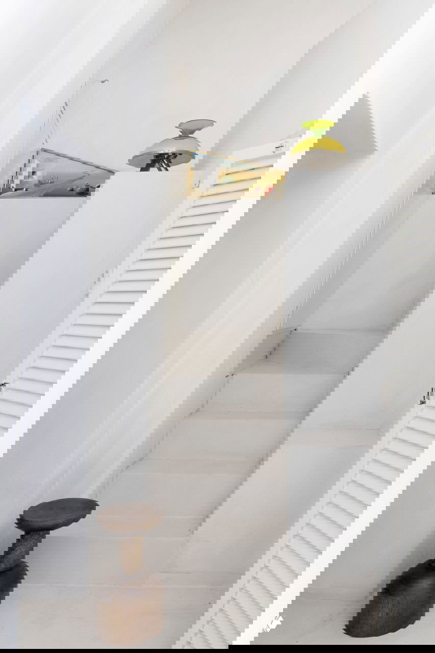 Узкая белая лестница и ниша в компактном доме: деревянный низкий табурет и декоративный элемент на фоне чистых стен и строгих форм.