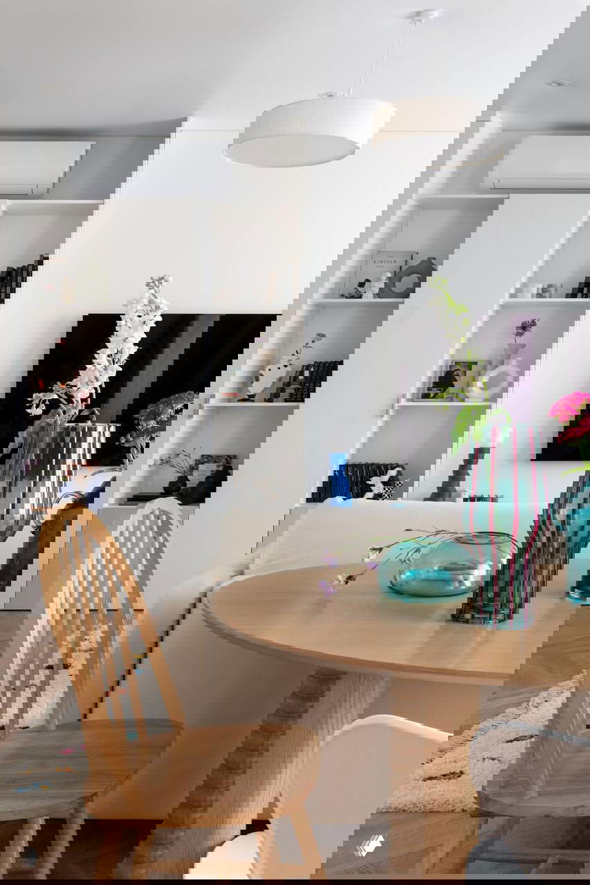 Обеденная зона и ТВ: круглый деревянный стол, стеклянные вазы бирюзового цвета и открытые полки с декором на светлом фоне.