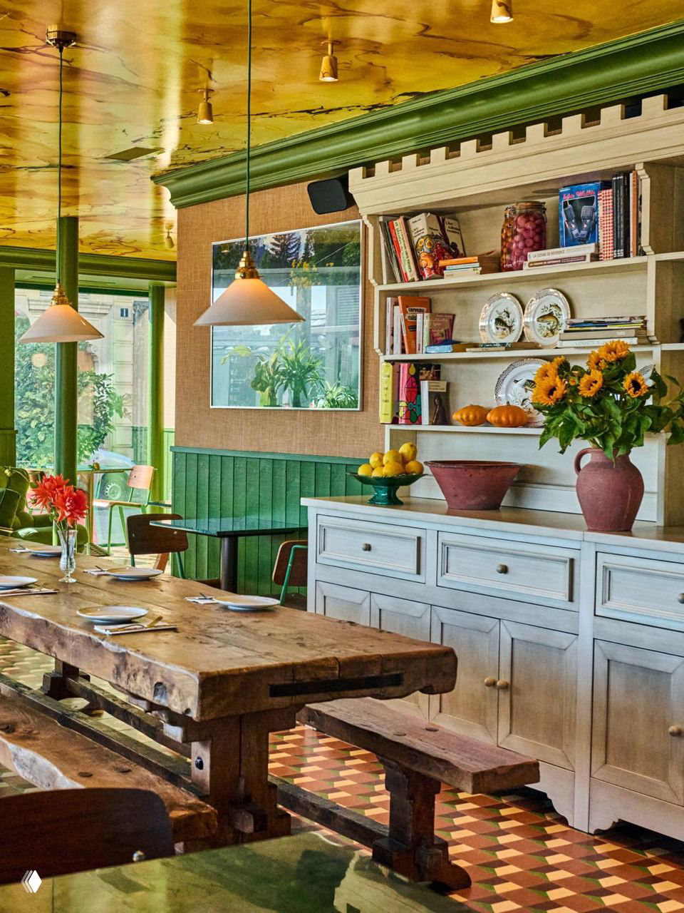 Интерьер ресторана с крупным деревянным столом, скамьями, сервировкой и декоративной тумбой, зеленые панели и желтый потолок.