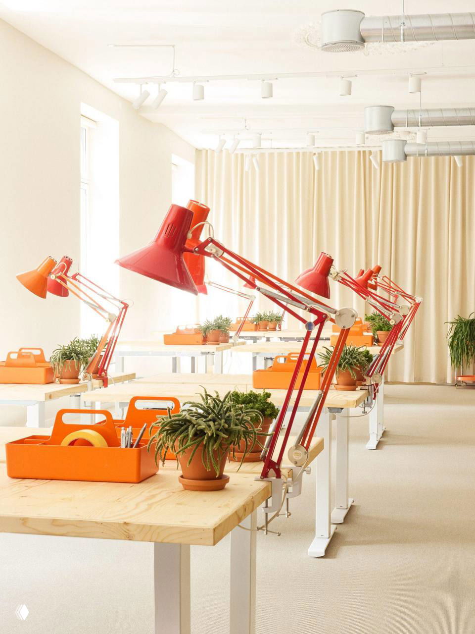 Рабочая зона офиса: длинные столы, красные лампы на струбцине, оранжевые органайзеры и зелёные комнатные растения для оживления пространства.