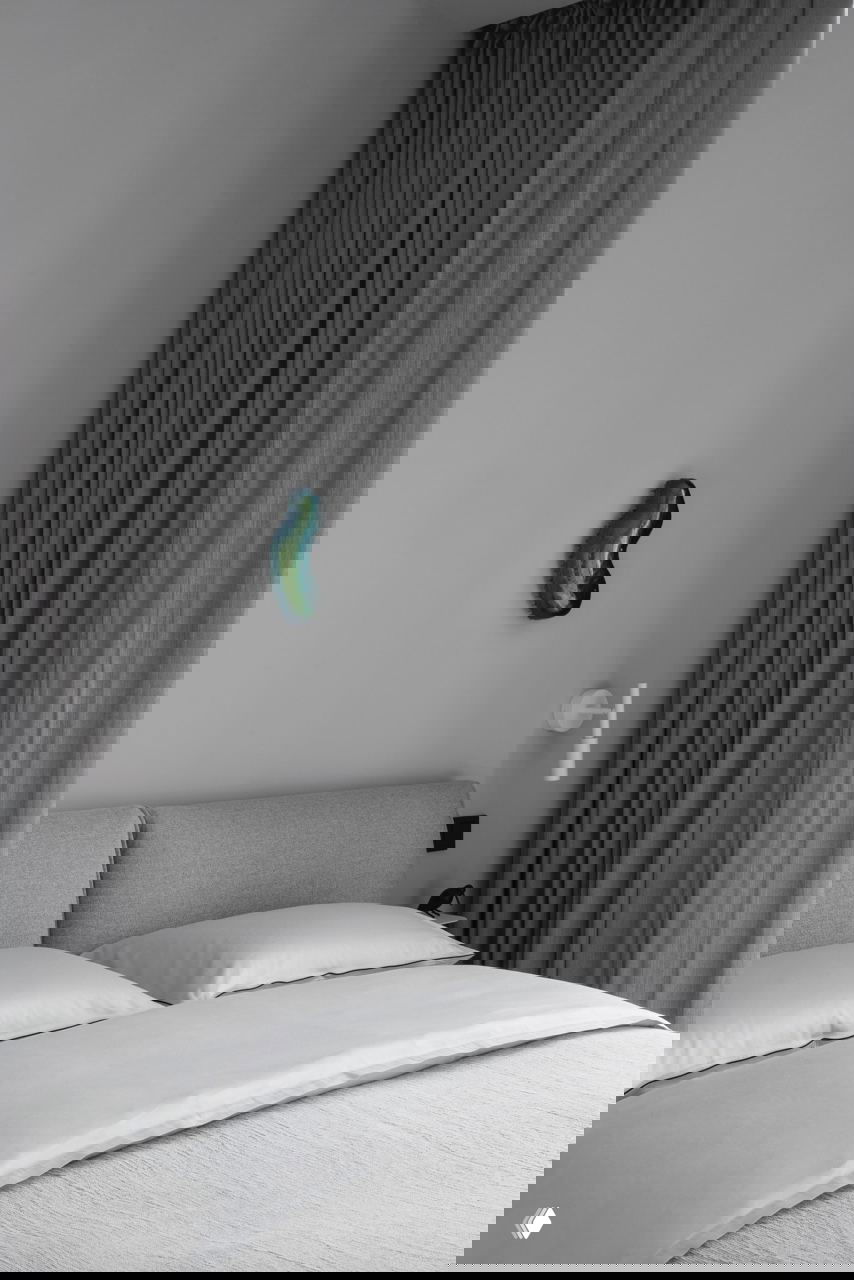 Спальня в приглушённых серых тонах: кровать с аккуратным текстилем, плотные шторы и небольшая цветная настенная скульптура как единственный яркий акцент.