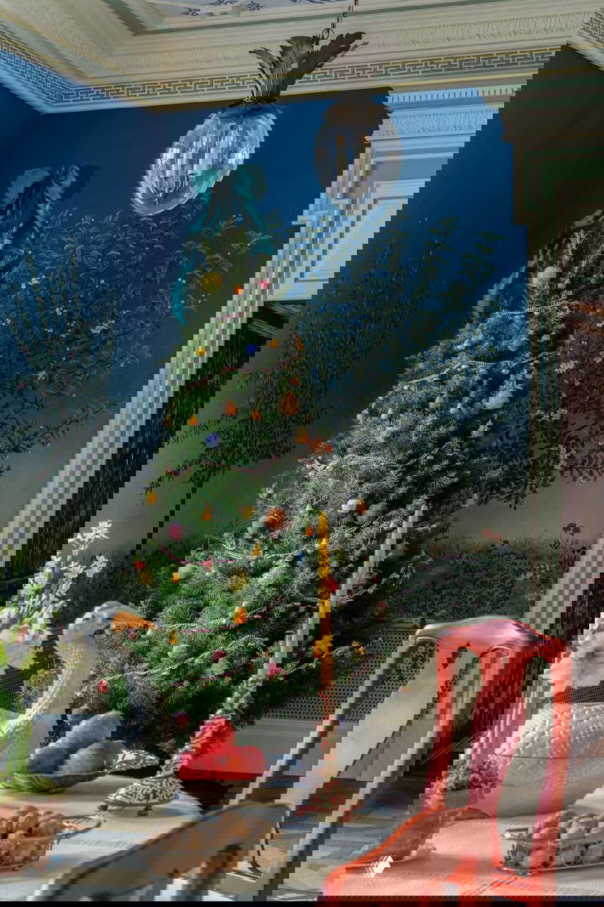 Столовая в фермерском доме с зелёной рождественской ёлкой на фоне синих стен и панорамного флористического панно, оформленного по эскизам Беаты Хойман.