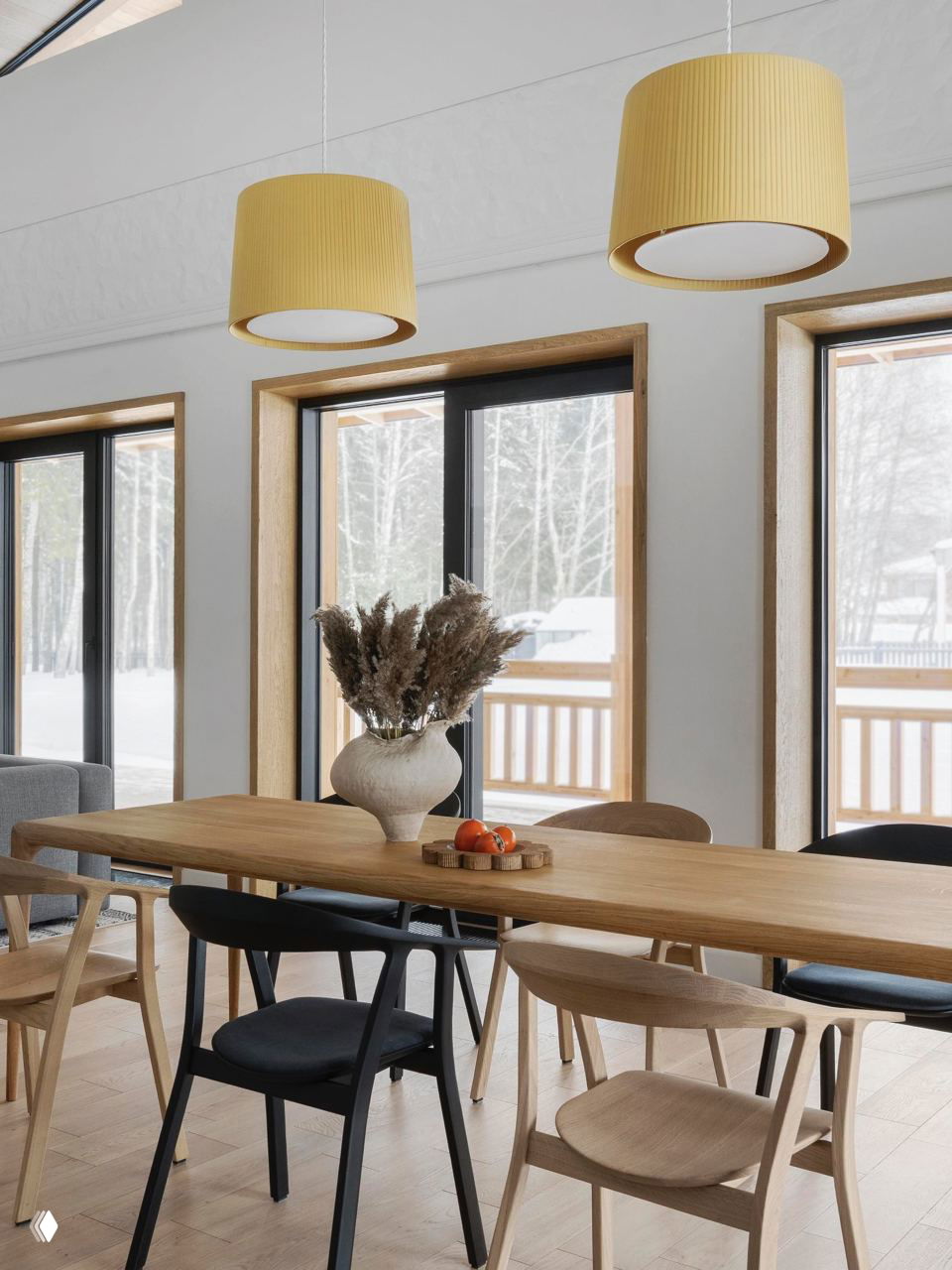Обеденная зона в скандинавском доме: длинный деревянный стол, стулья разной фактуры, желтые подвесные светильники и вид на заснеженный двор.
