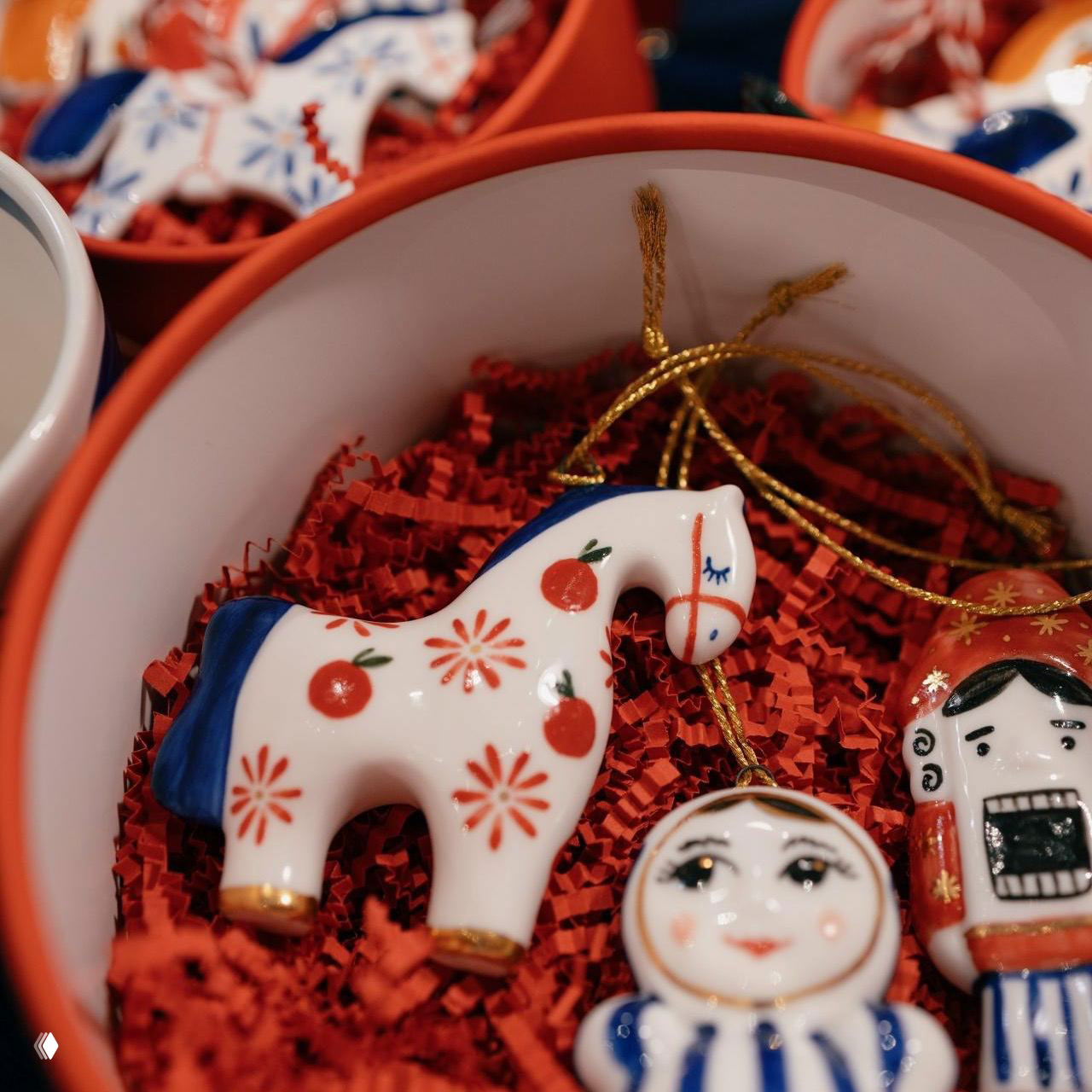 Небольшие керамические ёлочные игрушки — расписанная белая лошадка и мини‑матрёшка в подарочной коробочке с красной бумажной стружкой, предметы маркет‑предложения.