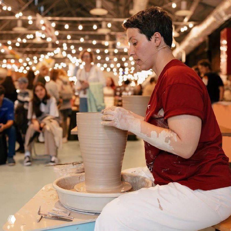 Фотография с визуальными артефактами: мастер за гончарным кругом и посетители на фоне праздничных гирлянд, кадр выглядит повреждённым цифровыми искажениями.