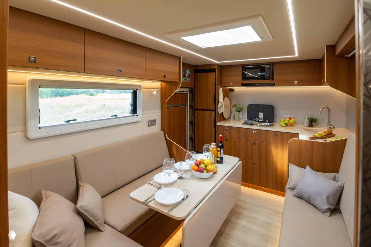 Интерьер автодома: столовая зона с раскладным столом, сервировкой, встроенной кухней и мягкими диванами, тёплая отделка деревом.