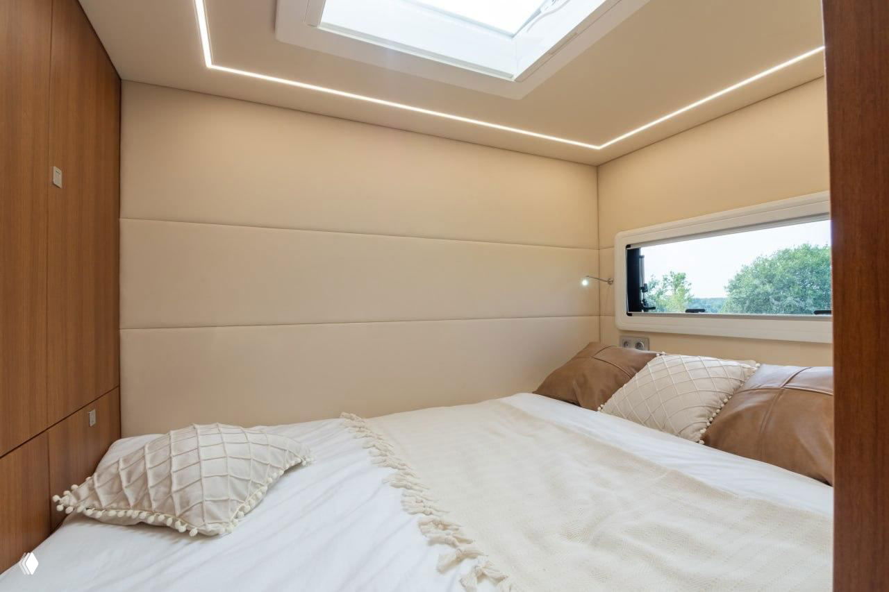 Главная спальня автодома с двуспальной кроватью, панорамным люком в потолке и светлой обивкой стен для комфортного отдыха в дороге.