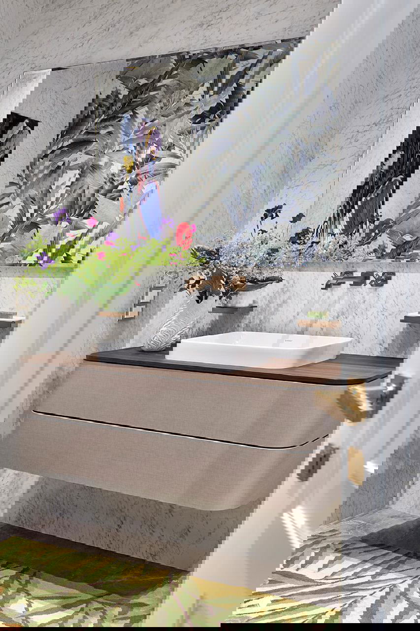 Ванная комната с мраморной отделкой, настенным зеркалом, плавающей тумбой с деревянной столешницей, латунным смесителем и выразительным растительным принтом на стене.