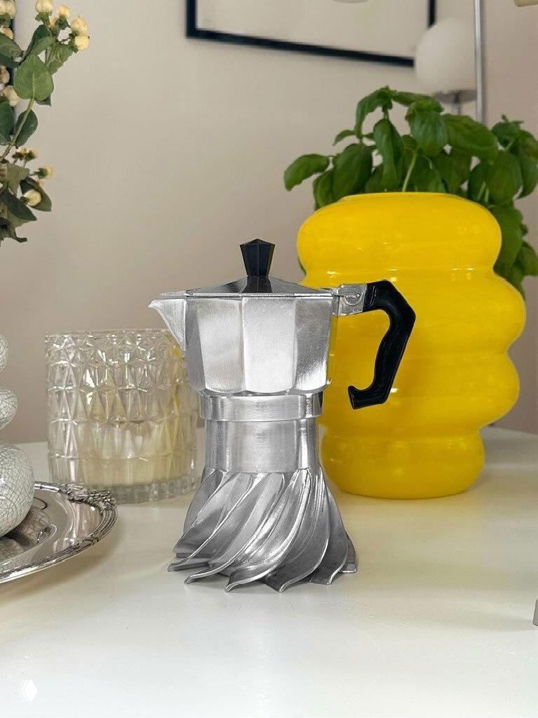 Фотография кофеварки Turbo Moka с турбинной спиральной основой на белом столе, на фоне желтой вазы и стеклянного подсвечника, вид спереди.