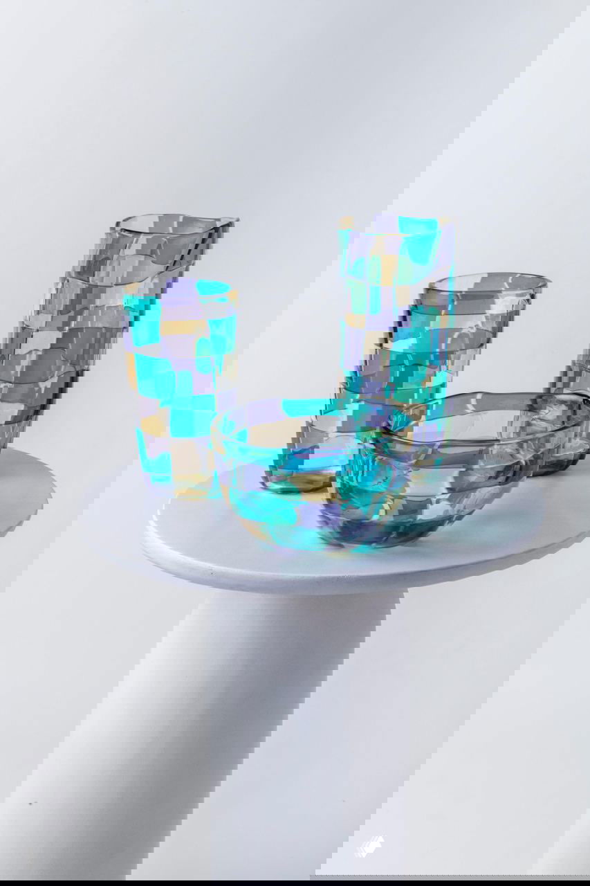 Три изделия коллекции Pezzati от Venini — две высокие вазы и глубокая чаша с мозаичным узором в желто-сиренево-мятной палитре на белом пьедестале.