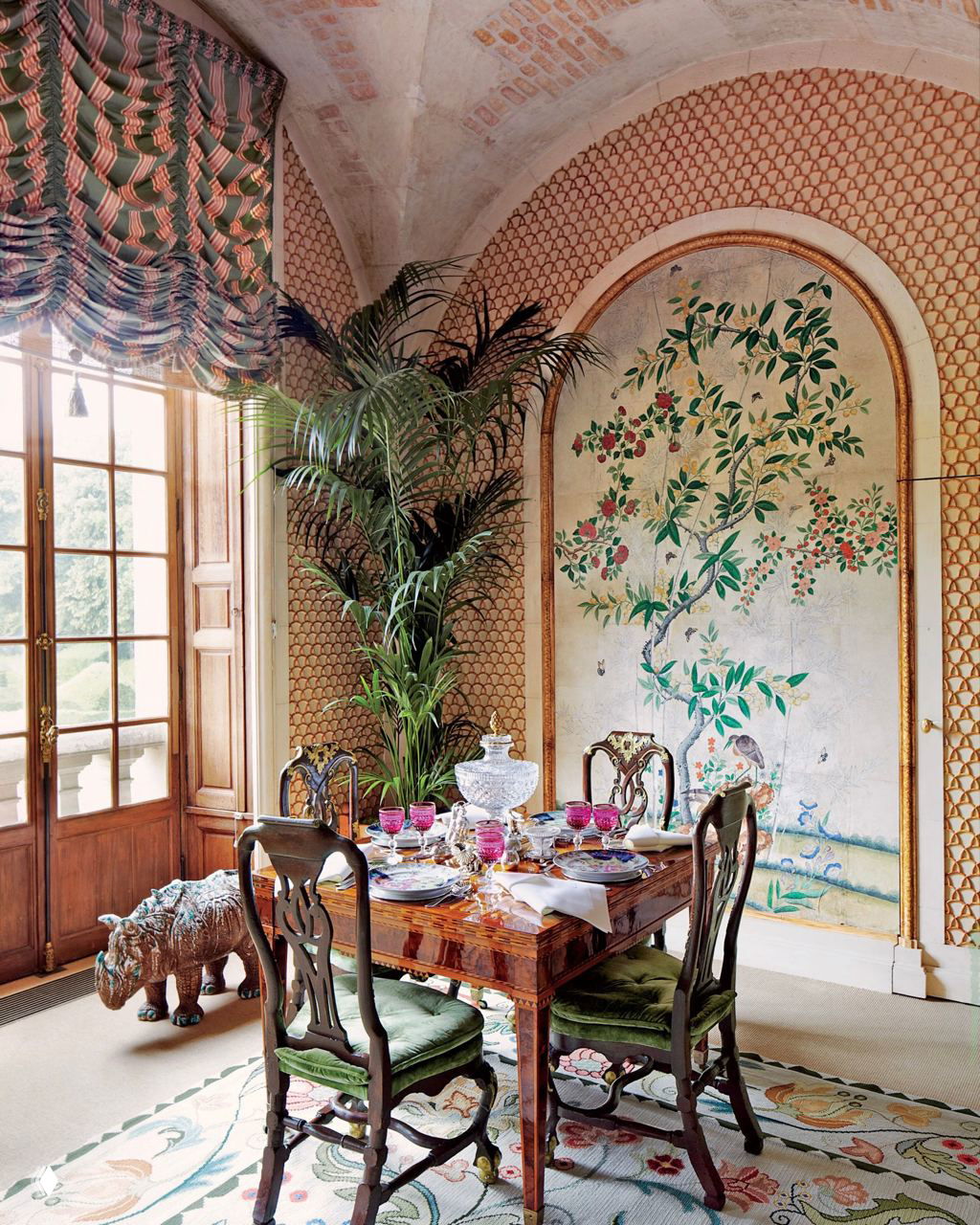 Столовая с арочными окнами, резным деревянным столом, сервировкой из фарфора и стеклянными розовыми бокалами, на фоне расписной панно.