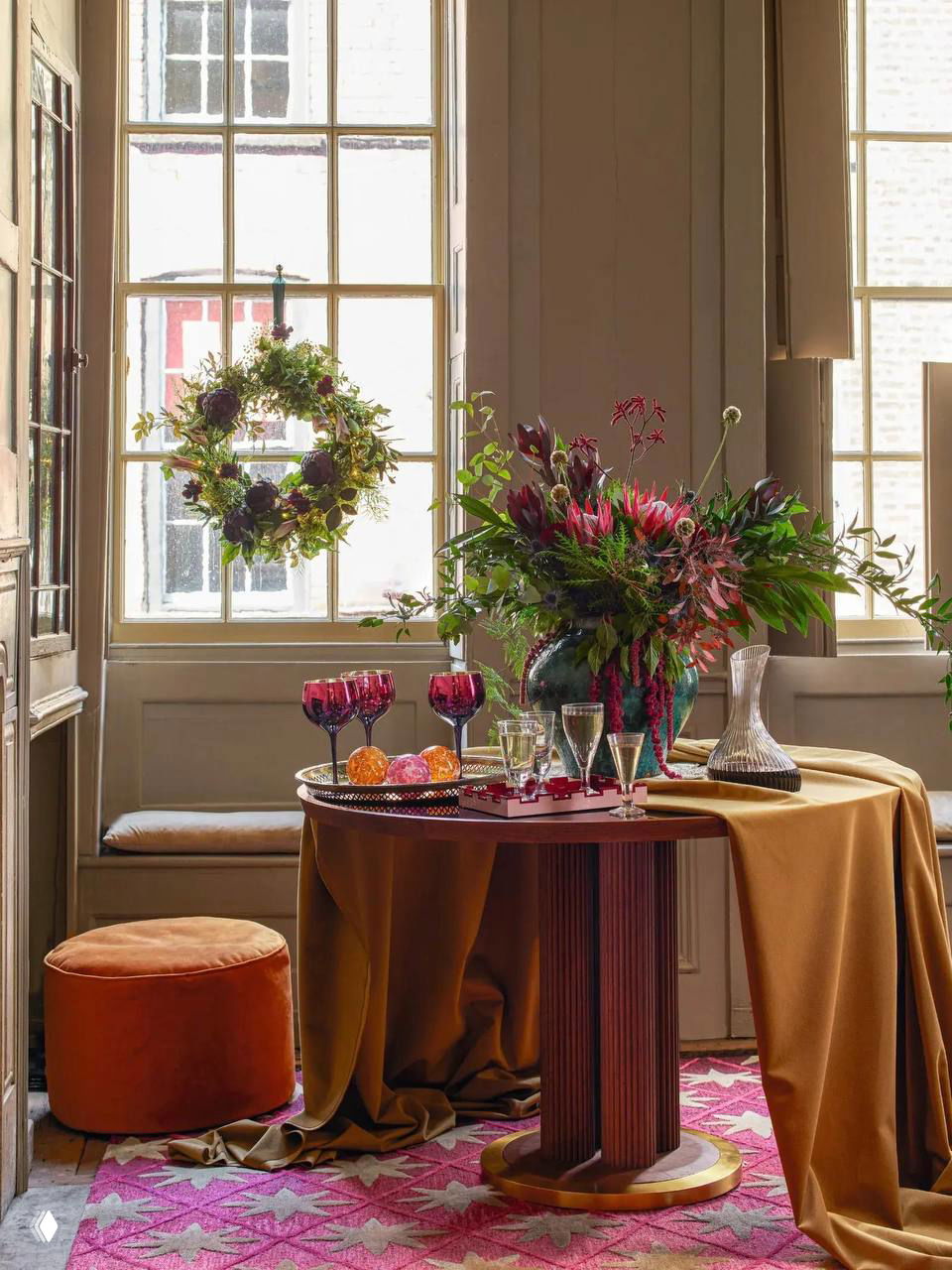 Праздничный уголок у окна: круглый стол с бархатной скатертью, пышный букет, фужеры и венок на стекле создают уютную новогоднюю сцену.