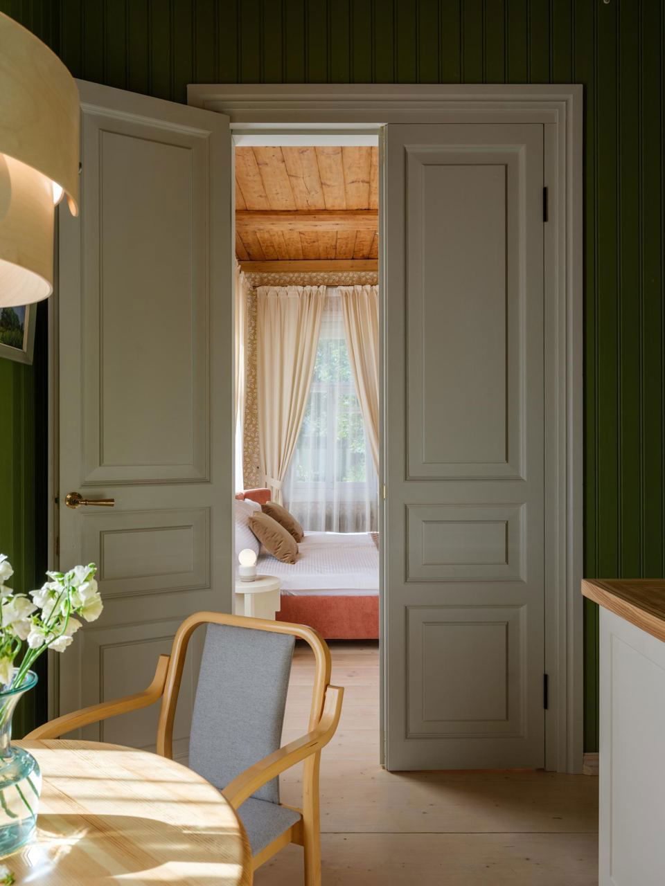 Проём между комнатами с открытыми двойными дверями, деревянный пол и мягкий дневной свет, вид на соседнюю спальню через дверной проём.