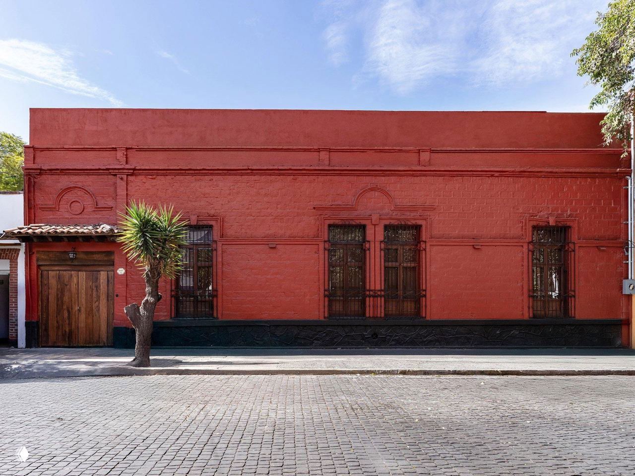 Фасад красного дома Casa Roja в Мехико: одноэтажное кирпичное здание с деревянной дверью, узкими окнами и деревом у тротуара, вид с улицы.