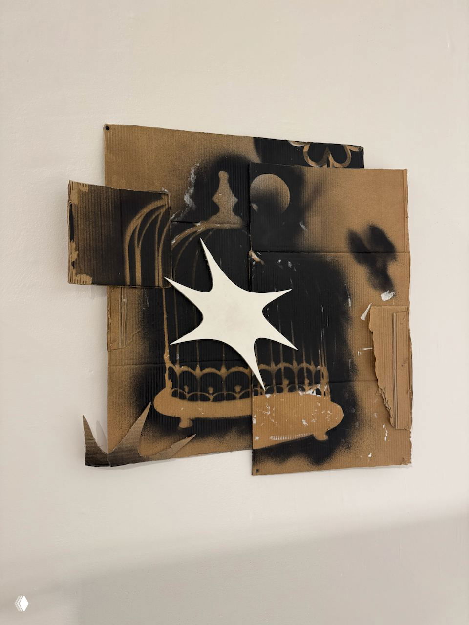 Коллаж из картона с чёрным аэрозольным рисунком и белым звездообразным элементом, размещённый на белой стене в выставочном зале
