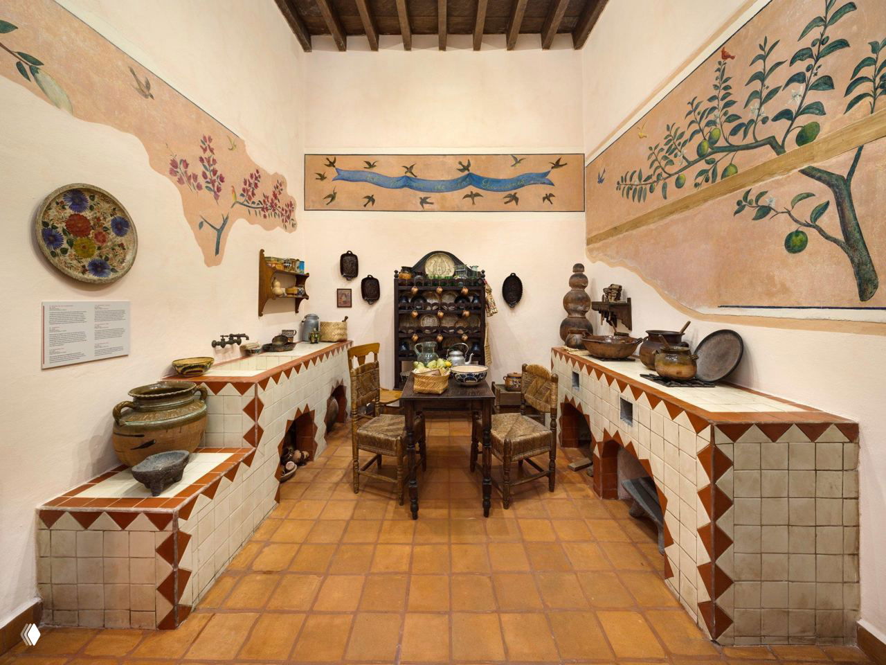 Воссозданная кухня Museo Casa Kahlo: плиточные рабочие поверхности, деревянная мебель и коллекция керамики на фоне фресок с растительными мотивами на стенах.
